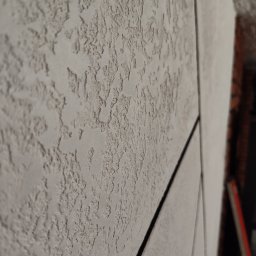 Gaga - Szara ściana z wyraźną fakturą, imitująca tynk strukturalny, z widocznymi liniami podziału na panele. Ujęcie z boku, podkreślające trójwymiarowość powierzchni.