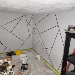 Gaga - Ściana z geometrycznym wzorem w trakcie malowania, widoczne narzędzia malarskie i taśma malarska. Fragment regału z drabiną w tle. Nietypowe wykończenie.