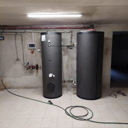 Gaga - Dwa pionowe, grafitowe zbiorniki akumulacyjne w kotłowni z szarą, surową ścianą i białą, kwadratową podłogą. Widoczne rury, zawory i przewody elektryczne.