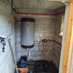 Gaga - Zamknięta przestrzeń z szarym zbiornikiem i czerwonymi rurami. Widoczne narzędzia i elementy instalacji. Surowe ściany z betonu.