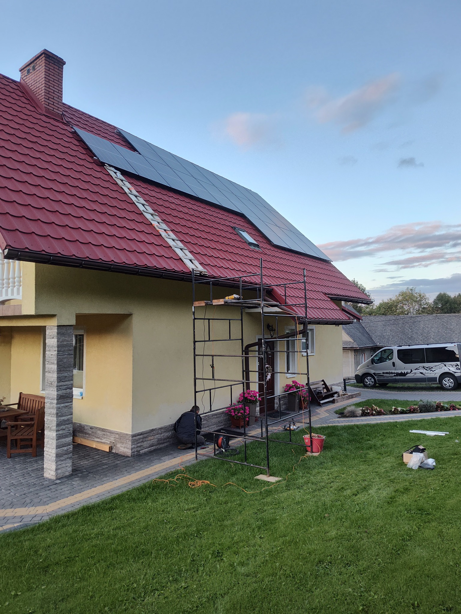 Montaż paneli fotowoltaicznych na dachu domu z czerwoną dachówką. Widoczne rusztowanie, drabina i częściowo sylwetka instalatora. Na trawniku narzędzia i akcesoria.