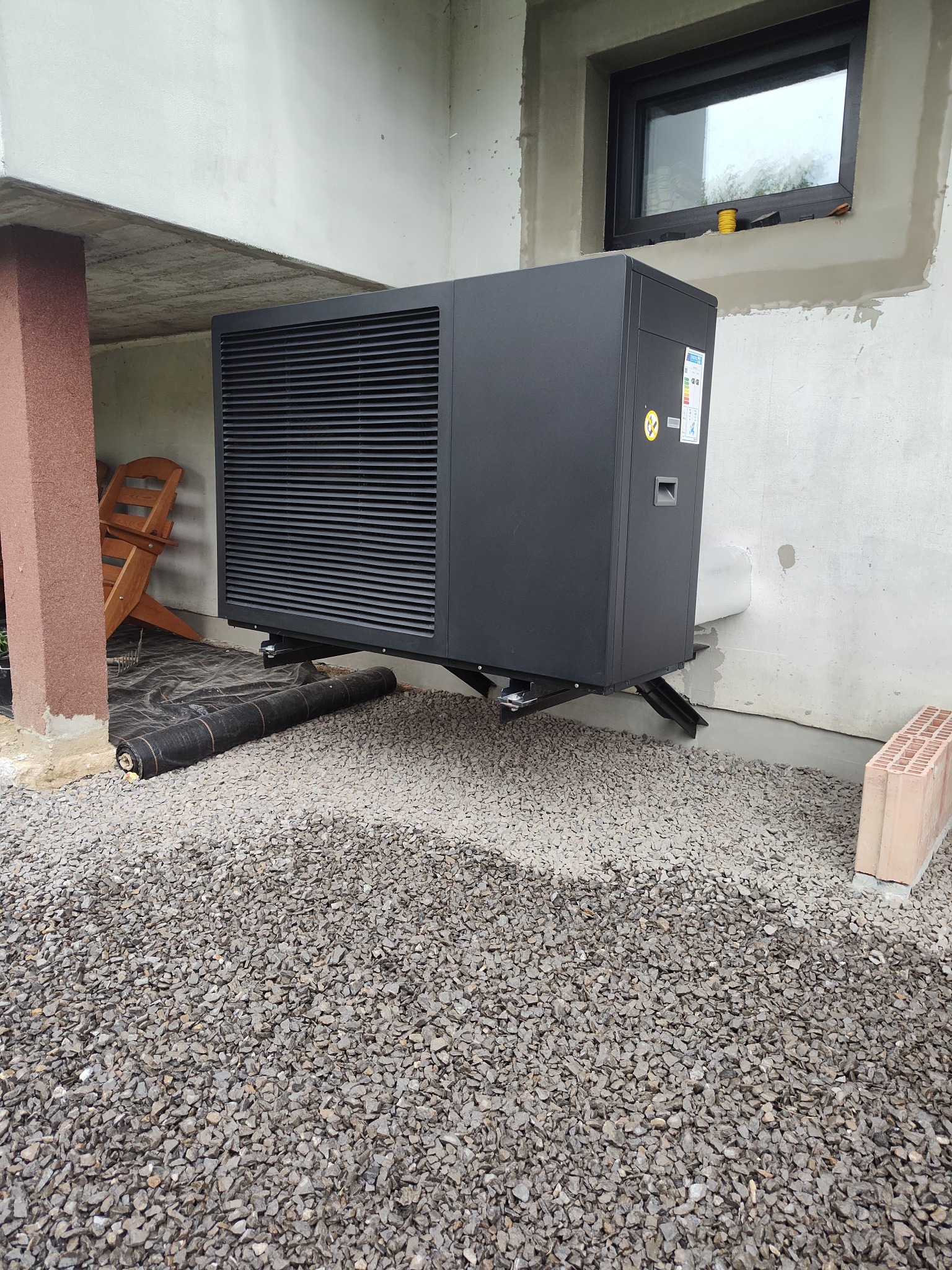 Czarna pompa ciepła na zewnątrz budynku, umieszczona na żwirowym podłożu, z widocznym fragmentem okna i elementami konstrukcyjnymi budynku w tle.