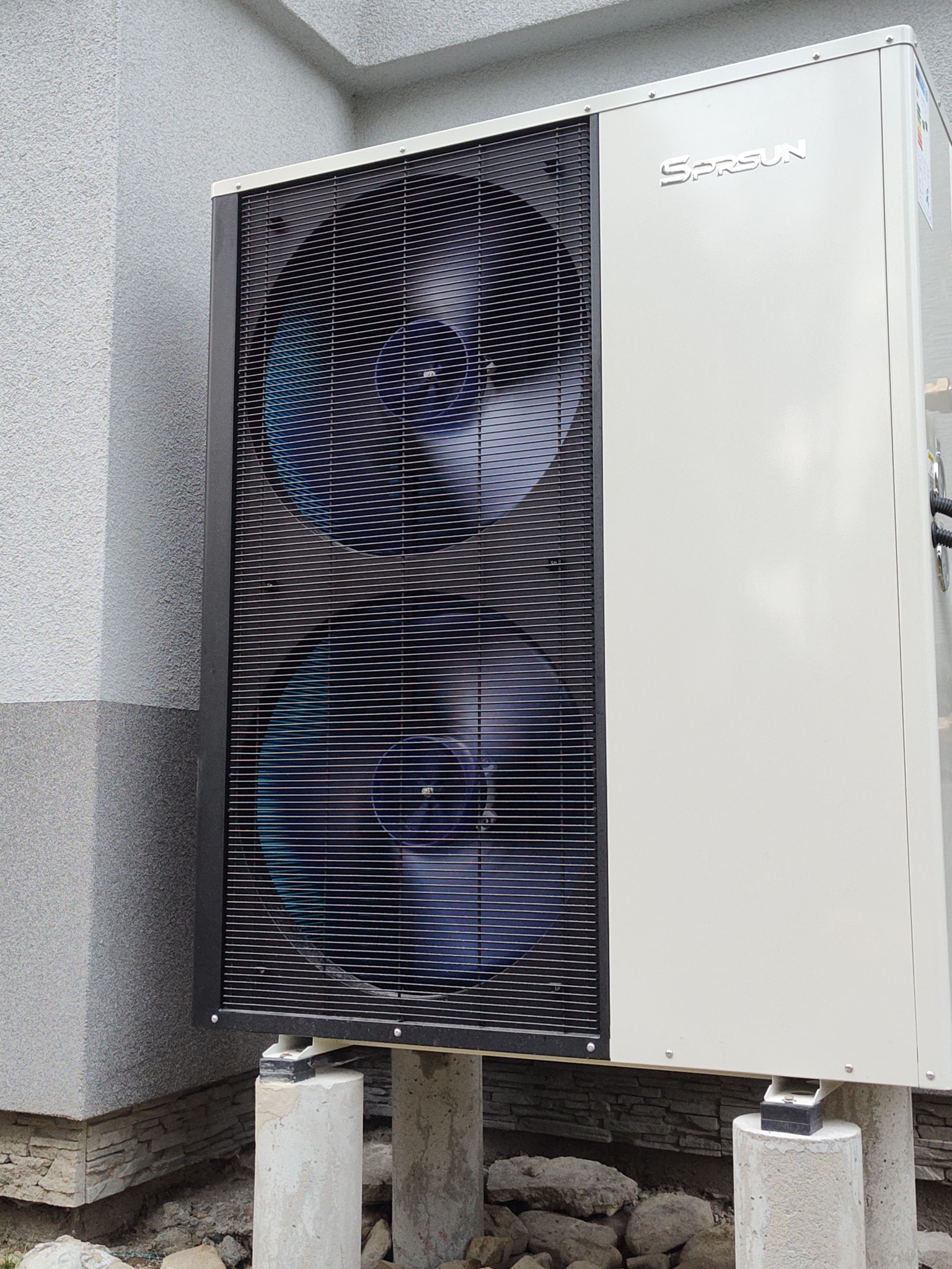 Biała pompa ciepła Sprsun z dwoma wentylatorami, zamontowana na betonowych słupkach przy szarej ścianie budynku z widocznym fragmentem elewacji.