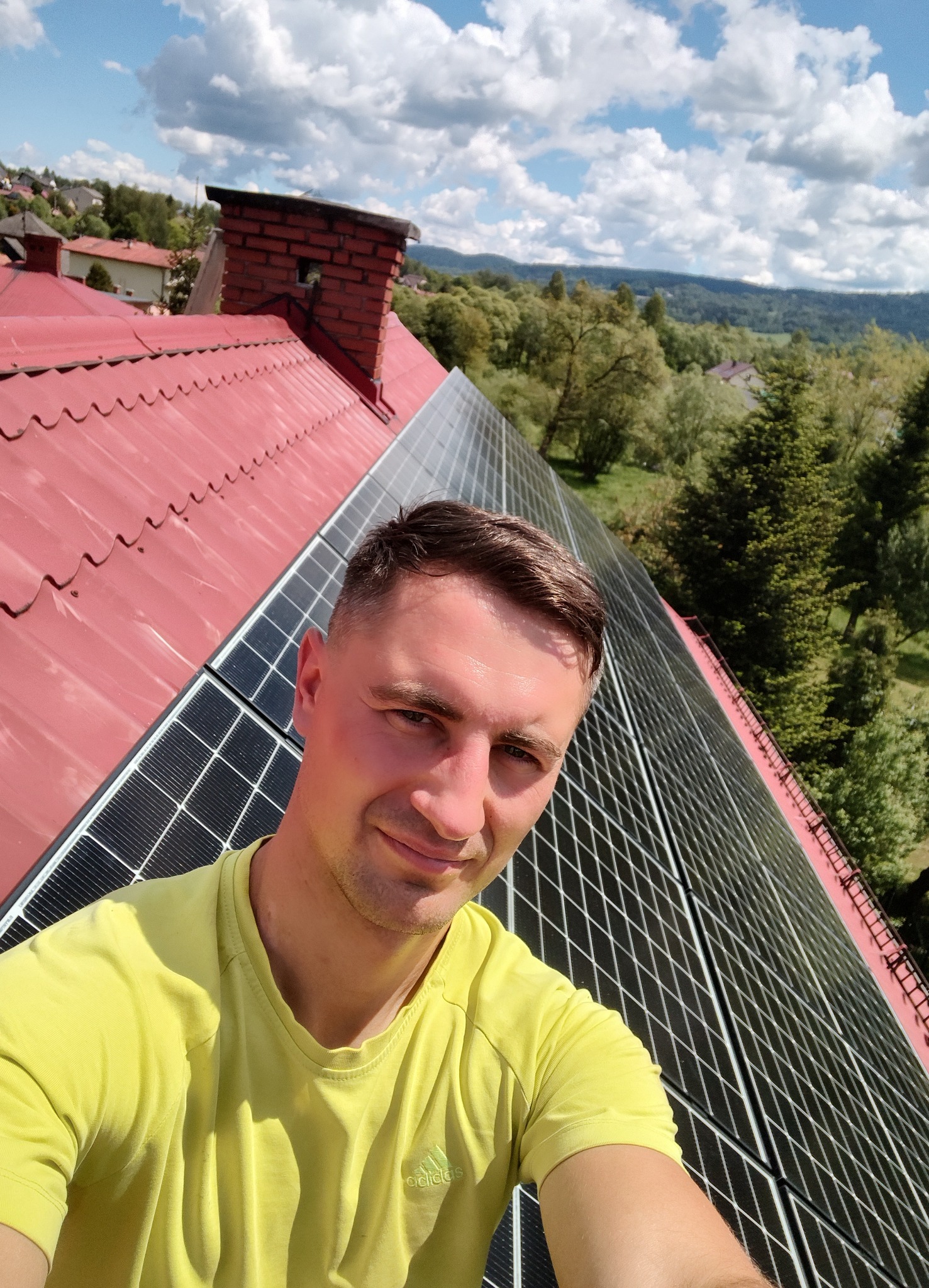 Instalator na dachu z panelami fotowoltaicznymi, widok z góry, w tle krajobraz zalesionych wzgórz i błękitne niebo z chmurami. Czerwona dachówka i ceglany komin.