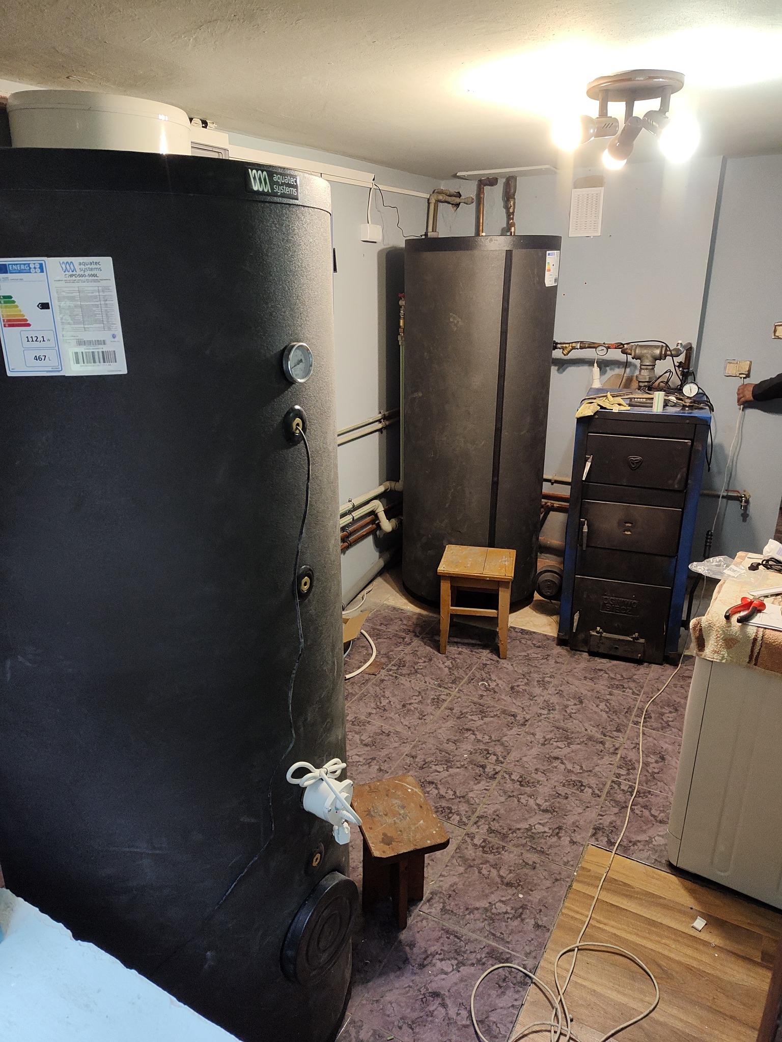 Dwa czarne zbiorniki akumulacyjne ciepła w kotłowni, widoczne rury i armatura, mały drewniany stołek i piec węglowy w tle, oświetlenie sufitowe.