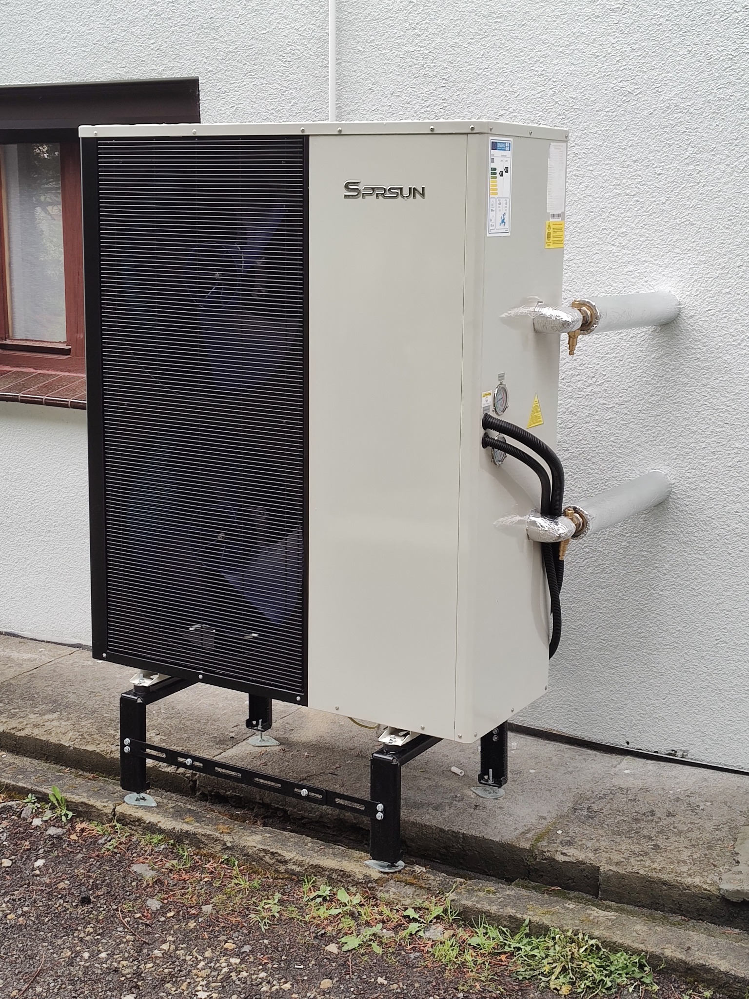 Zewnętrzna jednostka pompy ciepła SPRSUN zamontowana na czarnej, metalowej konstrukcji przy ścianie budynku z widocznymi rurami i oknem w tle.