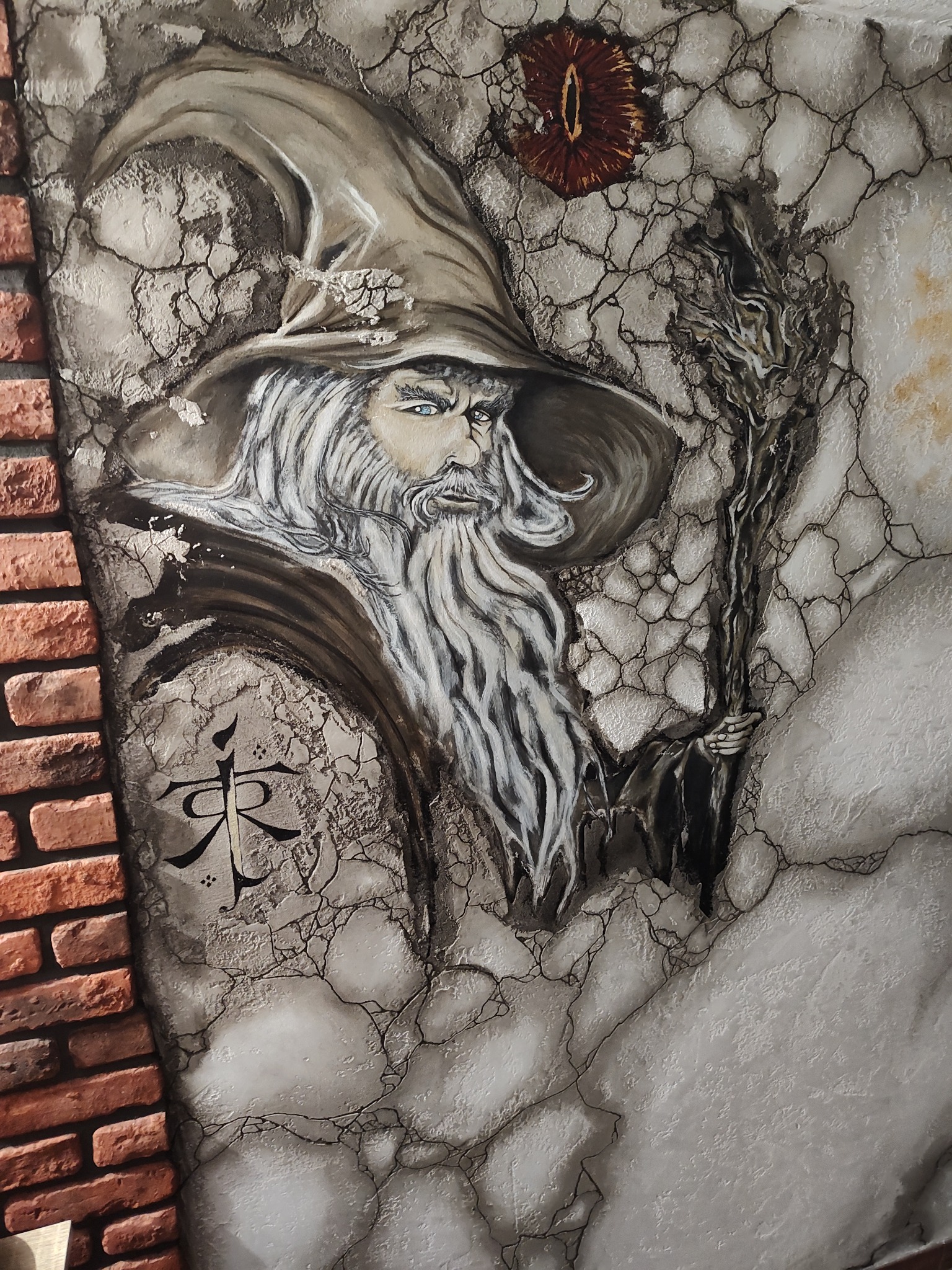 Artystyczne malowidło na ścianie z motywem czarodzieja w kapeluszu, z brodą i laską, obok fragmentu ściany z cegły. U góry oko Saurona. Styl fantasy, unikalna faktura.