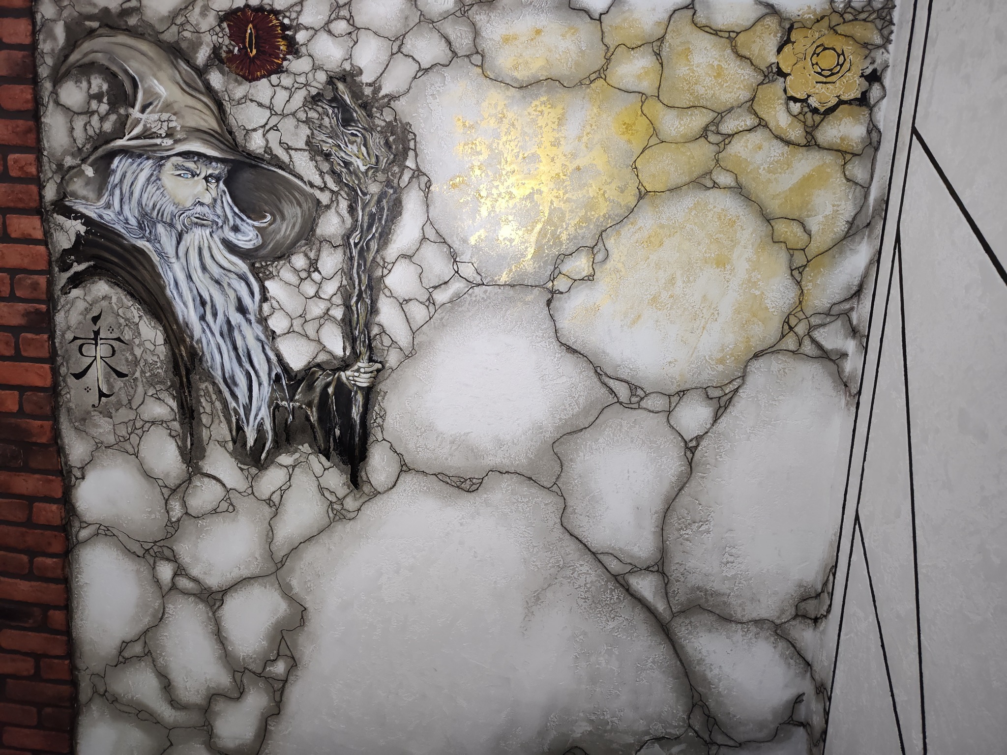 Artystyczne malowanie ściany z motywem czarodzieja i elementami dekoracyjnymi w odcieniach bieli, szarości i złota, imitujące pęknięcia i kamienną fakturę.