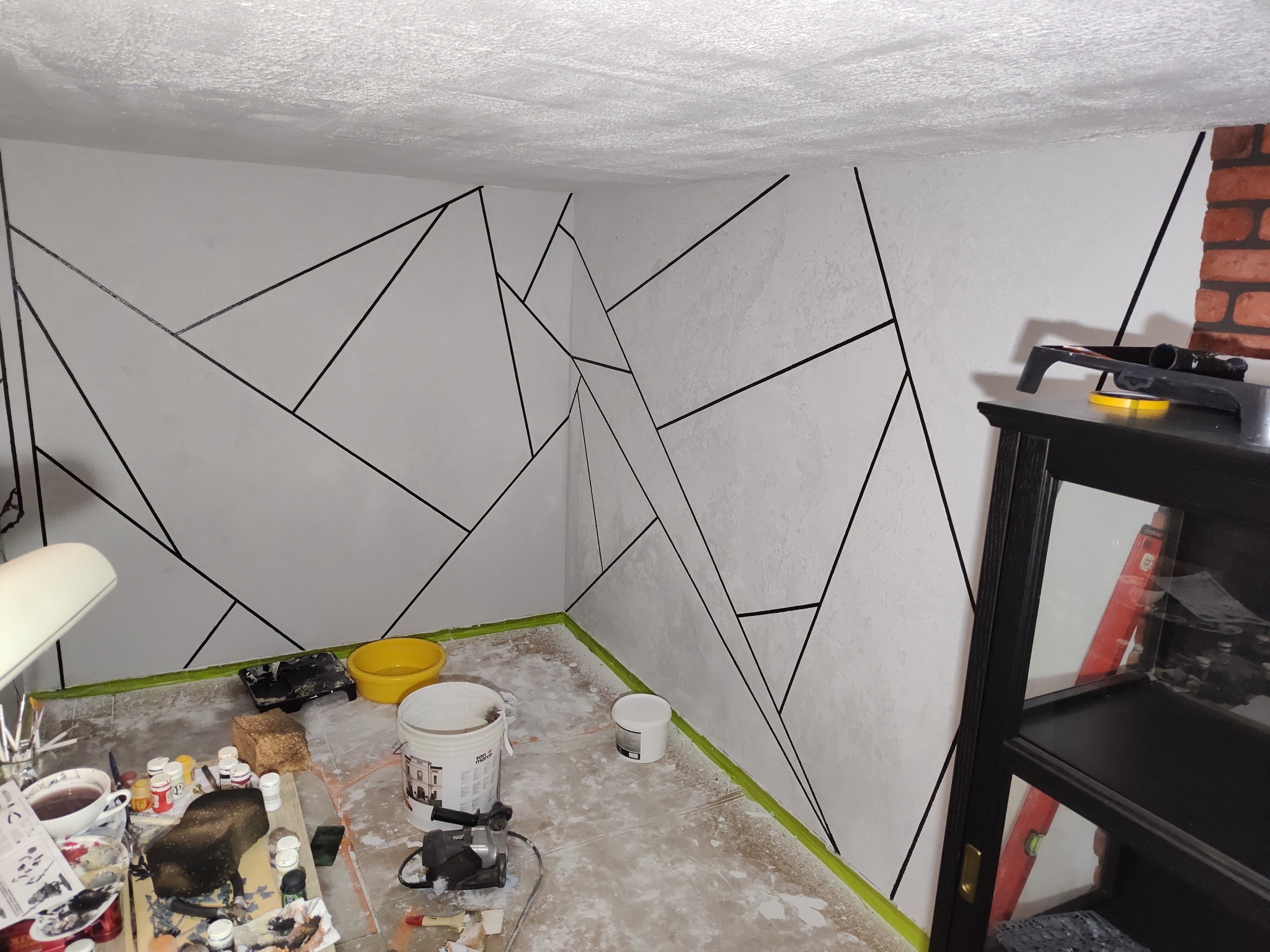 Ściana z geometrycznym wzorem w trakcie malowania, widoczne narzędzia malarskie i taśma malarska. Fragment regału z drabiną w tle. Nietypowe wykończenie.
