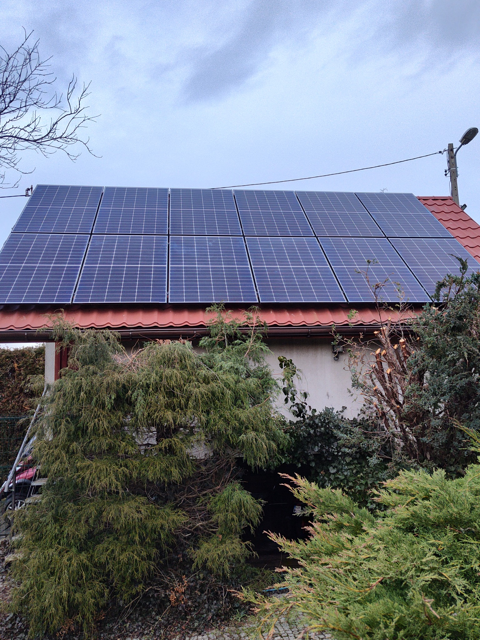 Panele słoneczne na dachu domu jednorodzinnego z czerwoną dachówką, widoczne z poziomu ogrodu, pochmurne niebo w tle. Częściowo zasłonięte przez zieleń.