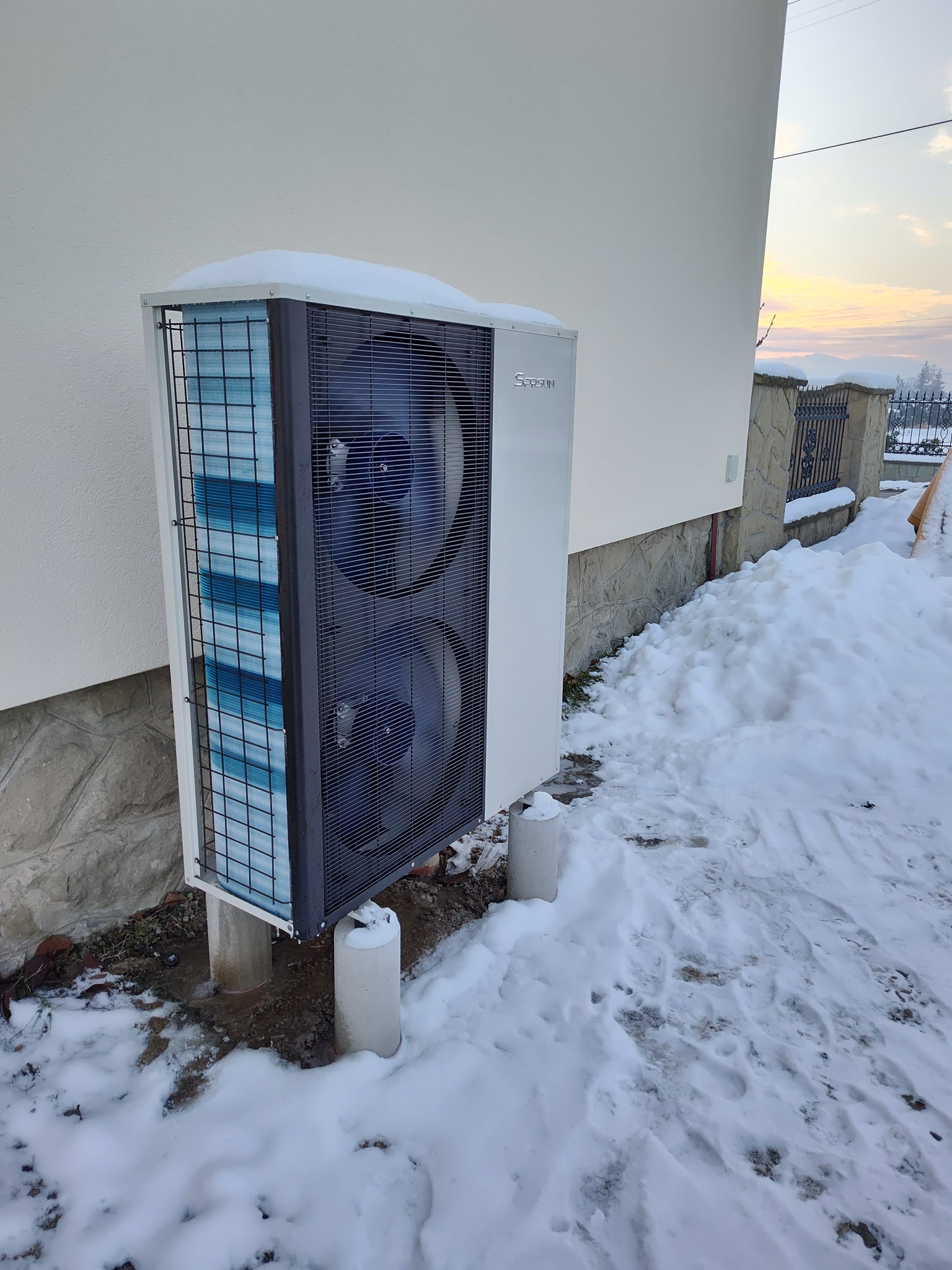 Biała pompa ciepła typu split, zamontowana przy budynku w zimowej scenerii. Na urządzeniu i wokół niego leży śnieg. Widoczne dwa wentylatory i metalowa kratka ochronna.