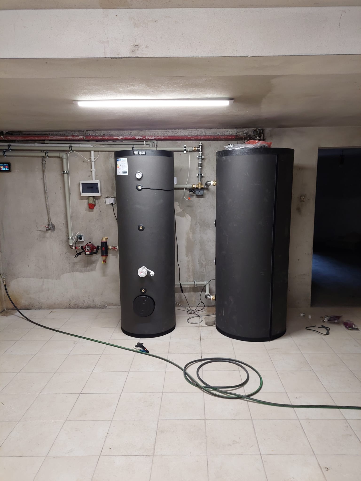 Dwa pionowe, grafitowe zbiorniki akumulacyjne w kotłowni z szarą, surową ścianą i białą, kwadratową podłogą. Widoczne rury, zawory i przewody elektryczne.