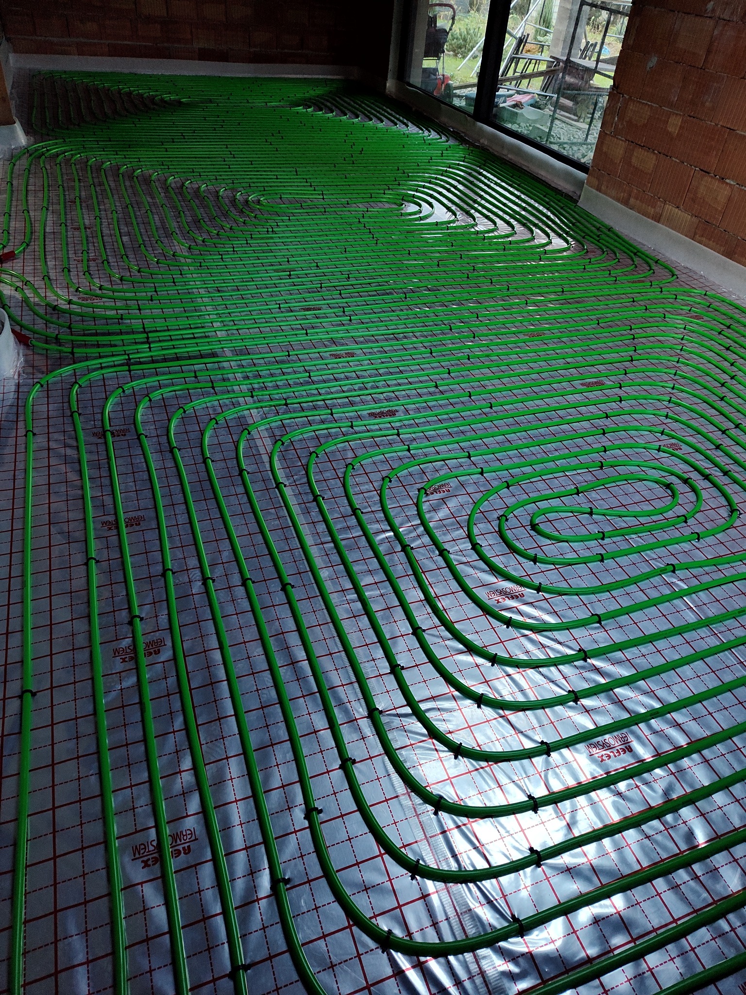 Zielone rury ogrzewania podłogowego ułożone spiralnie na folii z siatką, widok z góry, surowy stan przed wylewką. Instalacja w nowym budownictwie.