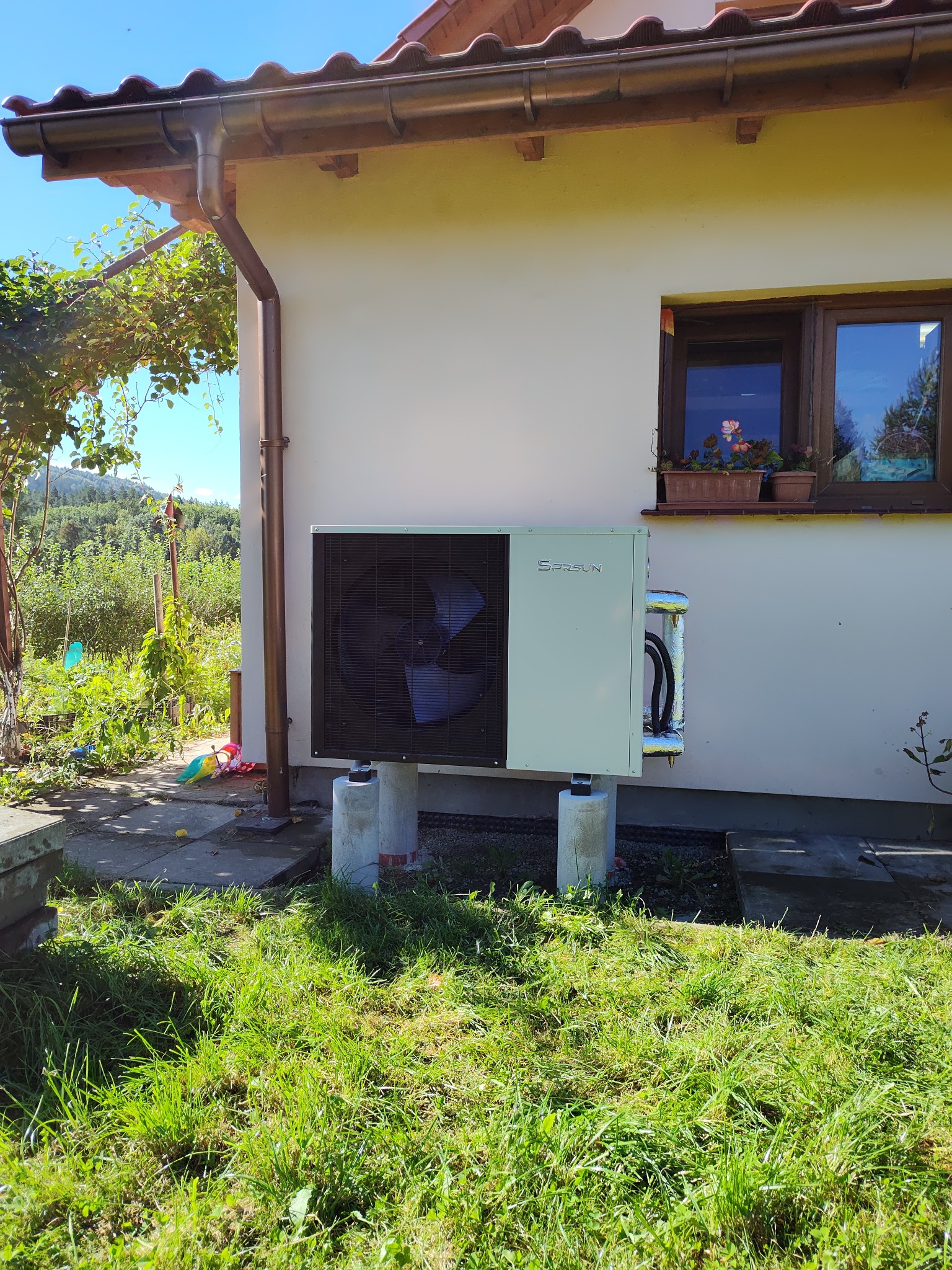 Biała pompa ciepła SPRSUN zamontowana na betonowych podstawach przy ścianie domu z oknem i kwiatami, na tle zielonego trawnika i drzew.