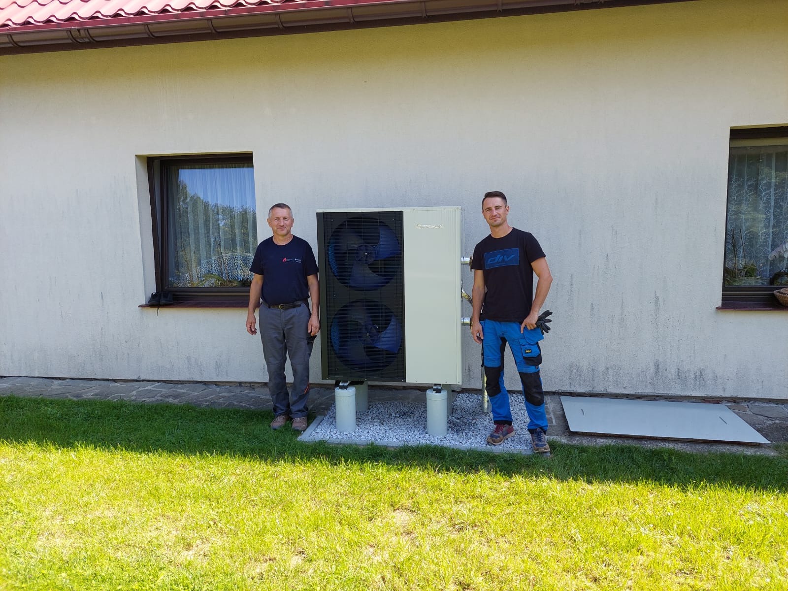 Dwaj instalatorzy stojący obok zainstalowanej pompy ciepła na zewnątrz budynku z oknem. Widoczne elementy instalacji i otoczenie budynku.