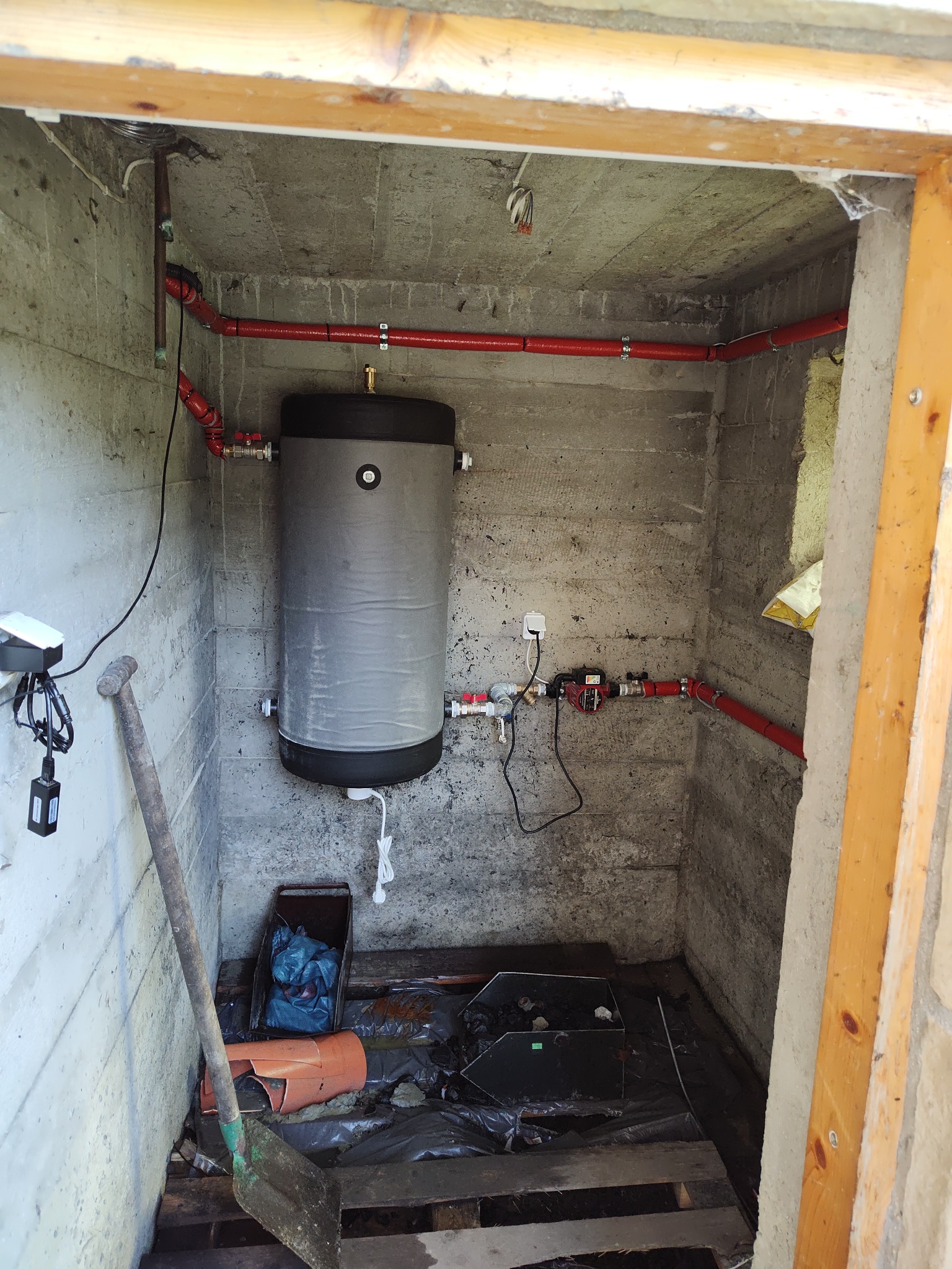 Zamknięta przestrzeń z szarym zbiornikiem i czerwonymi rurami. Widoczne narzędzia i elementy instalacji. Surowe ściany z betonu.