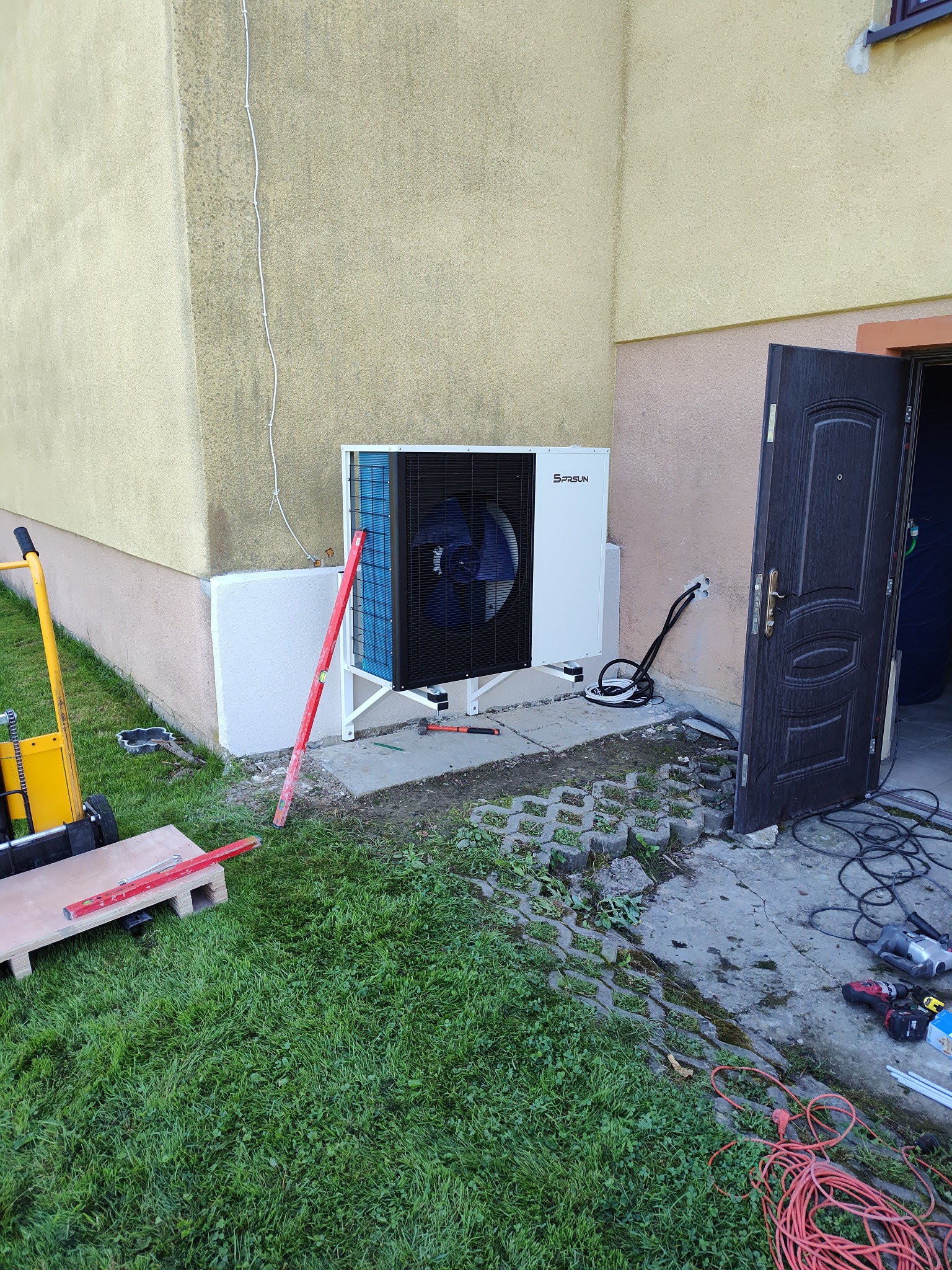 Biała pompa ciepła Sprsun zamontowana na elewacji budynku, widoczne narzędzia i poziomica, trawa i kostka brukowa wokół. Częściowo otwarte drzwi do garażu.