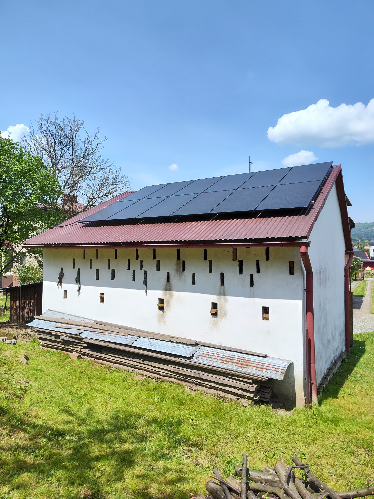 Biały budynek gospodarczy z panelami fotowoltaicznymi na czerwonym dachu. Stos blachy przy ścianie. Błękitne niebo z chmurami w tle. Widok z dołu.