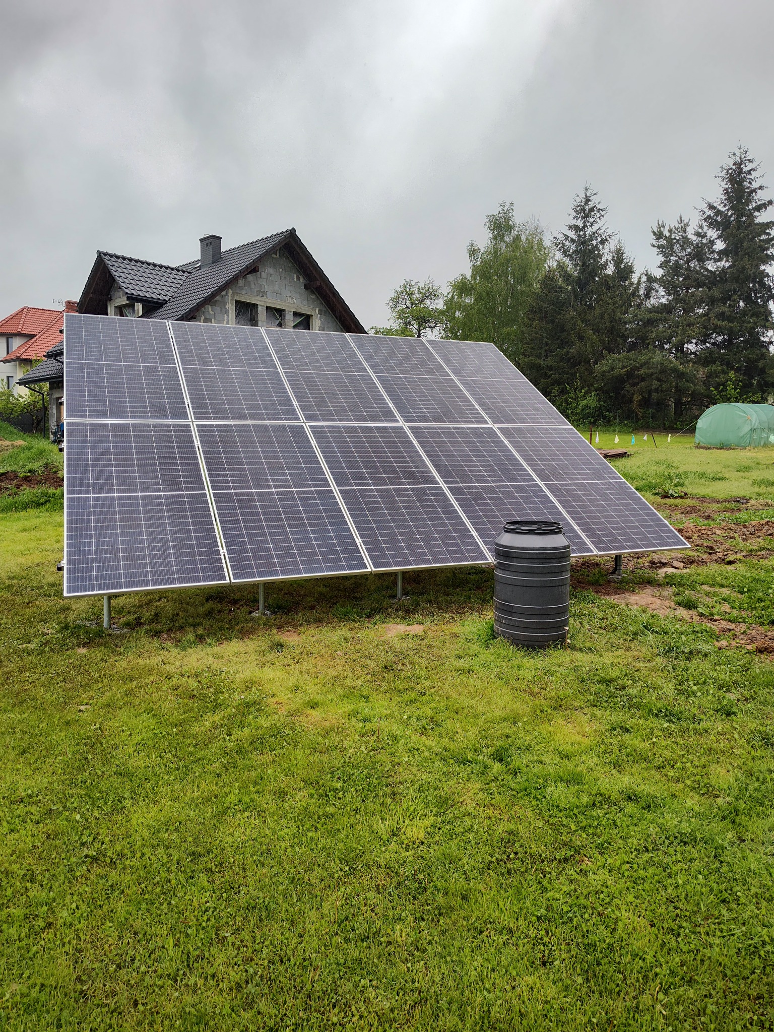Panel fotowoltaiczny na trawniku przed domem w budowie, obok czarny zbiornik na wodę. Pochmurne niebo w tle. Widoczne ślady prac ziemnych.