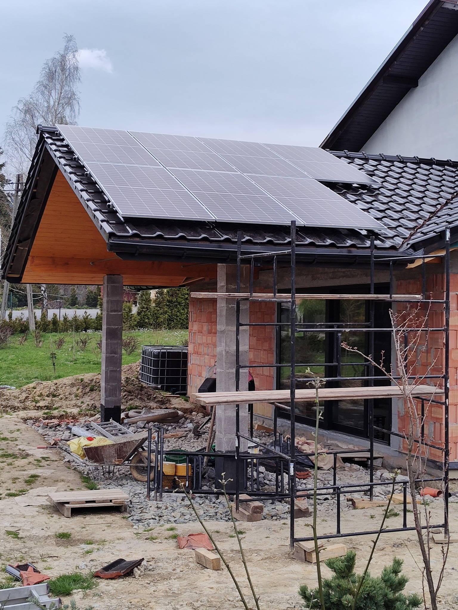 Montaż paneli fotowoltaicznych na dachu budynku w trakcie budowy. Widoczne rusztowania i materiały budowlane. Ujęcie z dołu, pochmurne niebo.
