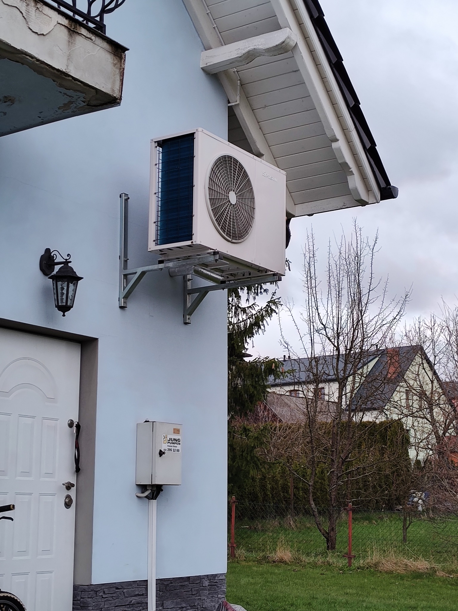 Biała pompa ciepła zamontowana na elewacji jasnoniebieskiego domu, widoczna lampa zewnętrzna i skrzynka elektryczna. W tle drzewa i domy.