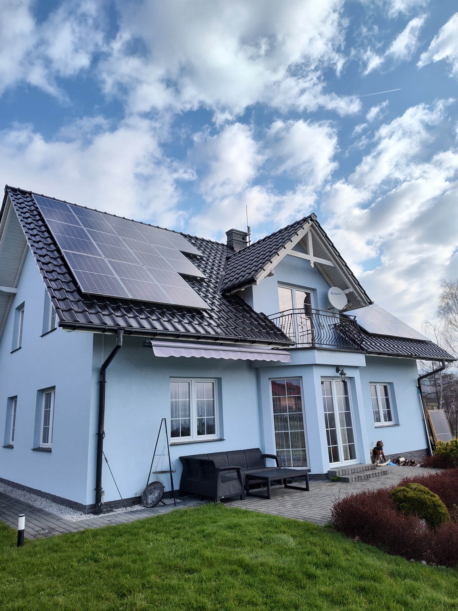 Jasny dom jednorodzinny z panelami słonecznymi na dachu, widok z ogrodu w słoneczny dzień. Na balkonie widoczna antena satelitarna.