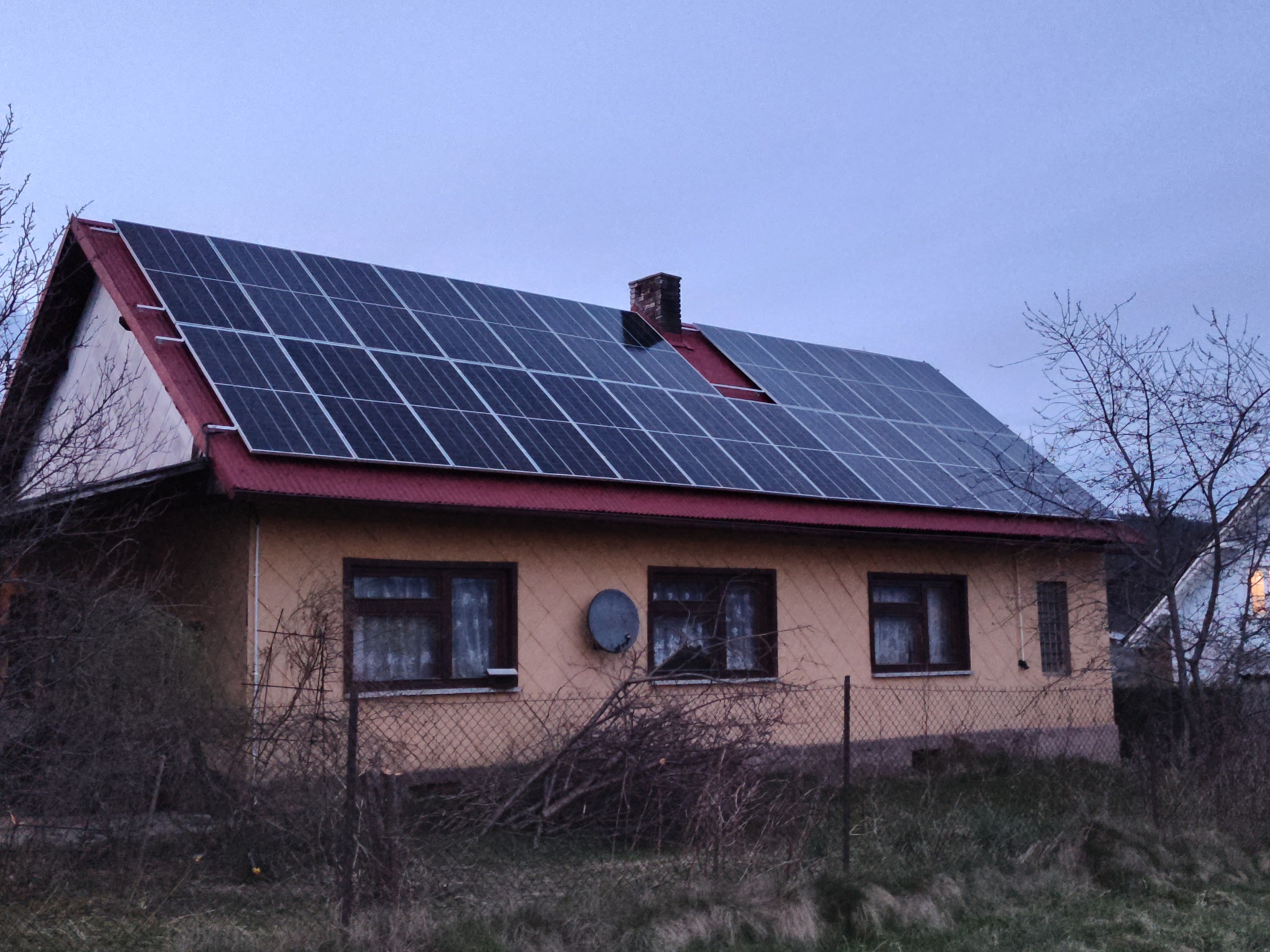 Dom jednorodzinny z panelami słonecznymi na dachu, widok z poziomu gruntu, w tle pochmurne niebo. Na ścianie budynku widoczna antena satelitarna.