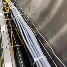 Karol Brożek - Uporządkowane kable elektryczne w metalowej korycie kablowej, zabezpieczone opaskami zaciskowymi. Widoczne kable w kolorach szarym, żółtym i czarnym.