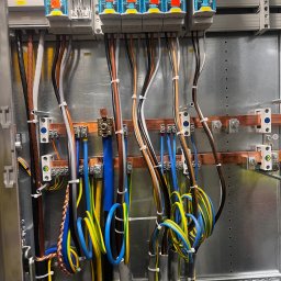 Karol Brożek - Rozdzielnia elektryczna z precyzyjnie ułożonymi przewodami, zabezpieczonymi niebieskimi modułami Wöhner i przymocowanymi do metalowej płyty z widocznymi punktami mocowania.