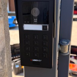 Karol Brożek - Czarny panel wideodomofonu Laskomex z klawiaturą numeryczną i czytnikiem RFID, zamontowany na szarym słupku bramy. Widoczny fragment kamiennego muru w tle.