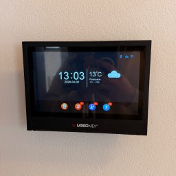 Karol Brożek - Panel sterowania Laskomex na ścianie, wyświetlający datę, godzinę, temperaturę i ikony powiadomień. Widoczny refleks światła na ekranie. Minimalistyczny design.