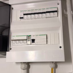 Karol Brożek - Rozdzielnica elektryczna Schneider z bezpiecznikami B16 i C16, widoczne przewody. Otwarta obudowa ukazuje wnętrze instalacji. Biała ściana w tle.