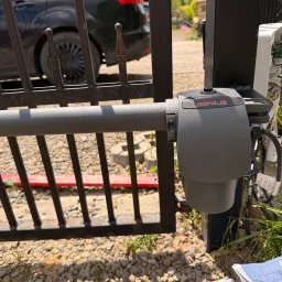 Karol Brożek - Zamontowany siłownik Genius do bramy wjazdowej, widoczna elektronika w obudowie, brama metalowa z elementami rdzy, tło z kamieni i trawy.