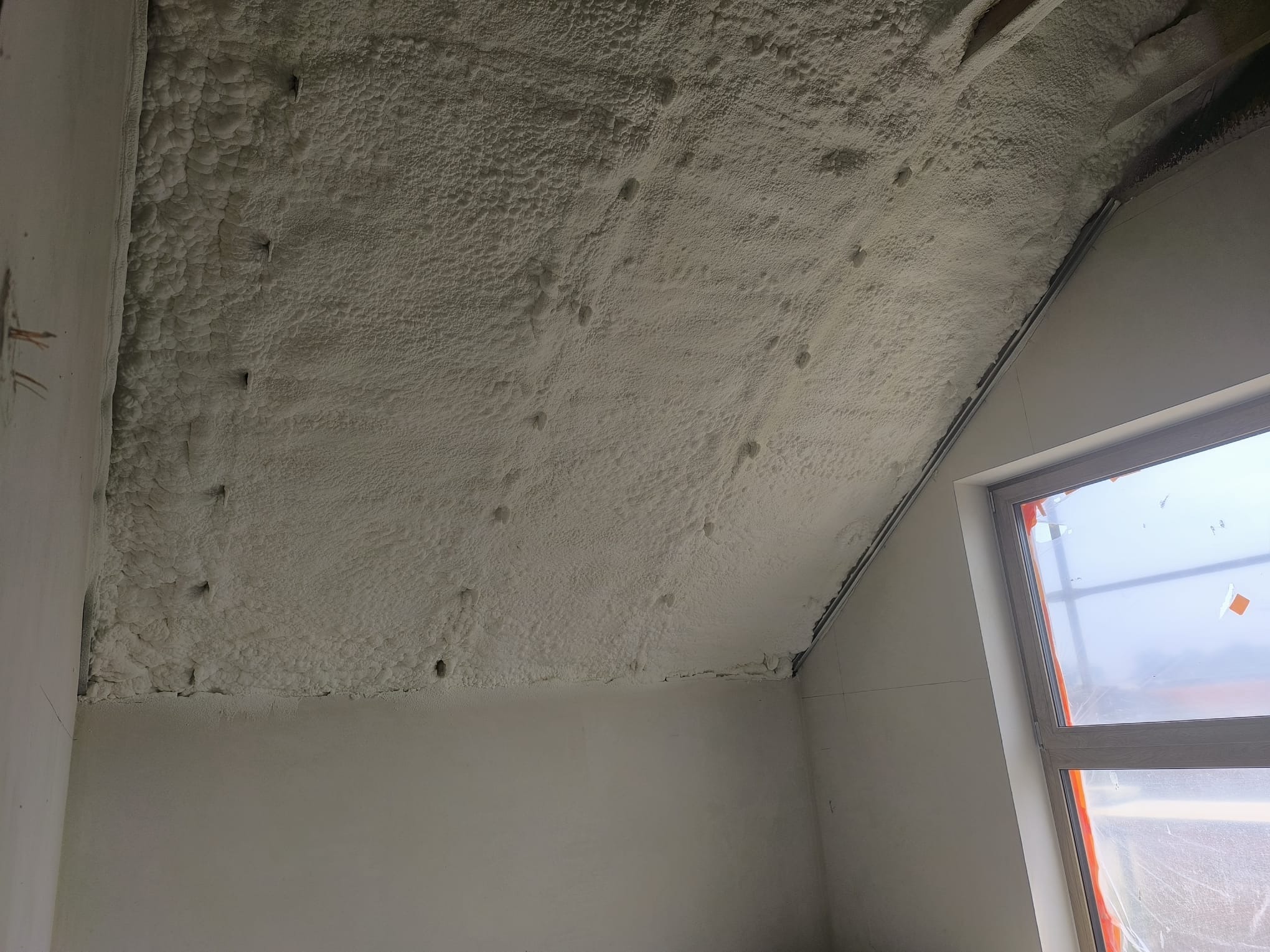 Poddasze po ociepleniu pianką PUR, widoczne nierównomierne pokrycie pianką, okno dachowe z folią ochronną. Detal wykończenia izolacji.