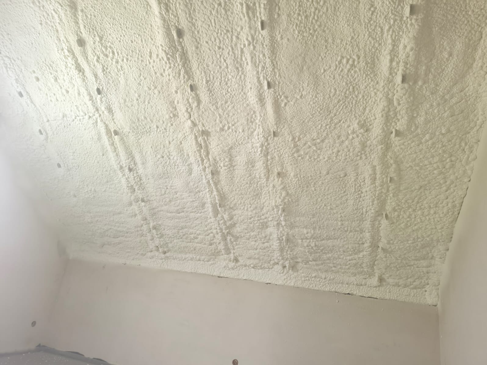 Poddasze po ociepleniu pianką PUR, widoczne równomierne pokrycie izolacją, z detalami faktury piany i wystającymi elementami montażowymi. Jasne, neutralne kolory.
