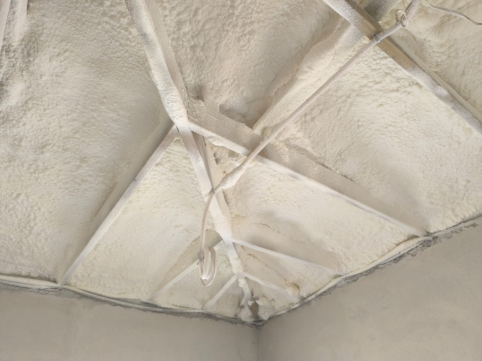 Poddasze z widoczną strukturą pianki izolacyjnej, pokrywającą krokwie i elementy konstrukcyjne. Szorstka faktura piany w jasnym odcieniu kremu. Widok z dołu.