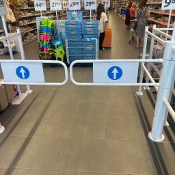 Neco PIOTR BINKOWSKI - Biała barierka z niebieską strzałką w supermarkecie, ograniczająca dostęp do alejek. W tle regały z towarami i klienci.