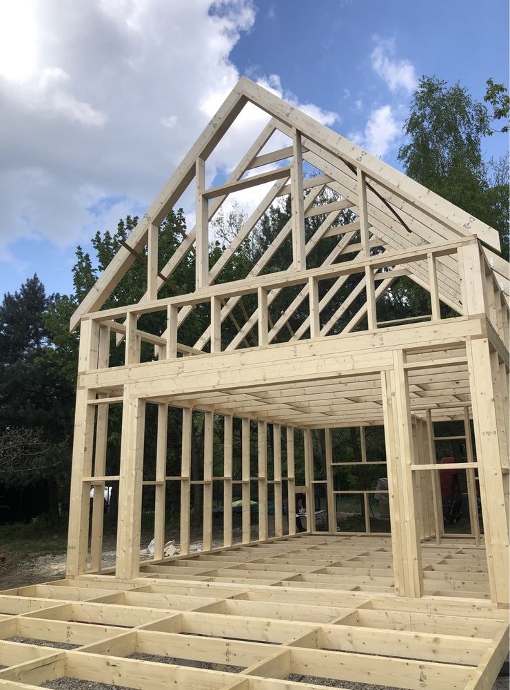 Szkielet domu w trakcie budowy: drewniana konstrukcja z widocznym dachem, ścianami i podłogą na tle zieleni i błękitnego nieba z chmurami. Widok z dołu.