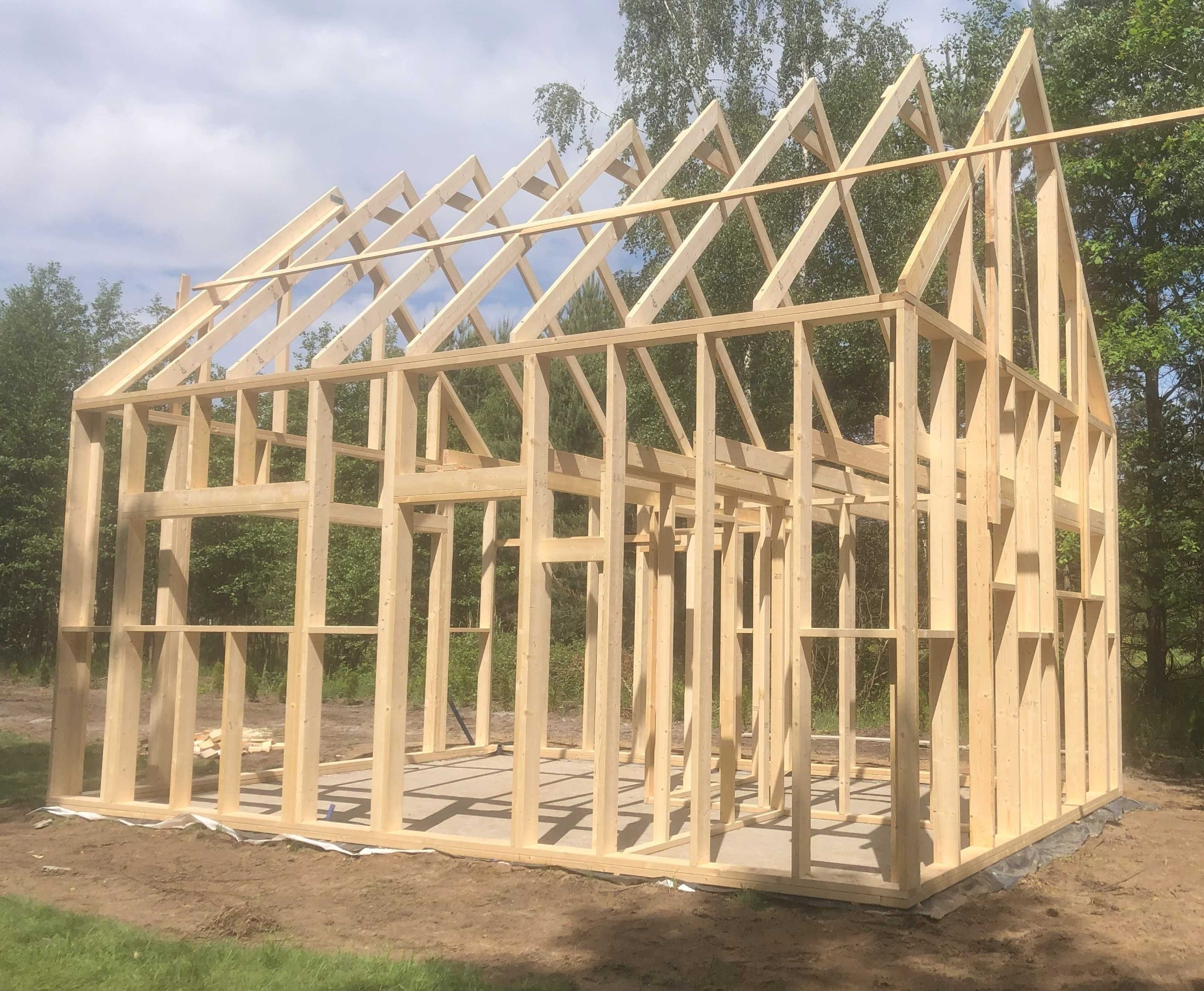 Konstrukcja szkieletowa domu w trakcie budowy, widoczne drewniane ramy ścian i dachu na betonowej płycie fundamentowej, z zielonym tłem drzew.