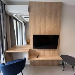 CONNECT MEBLE - Nowoczesna zabudowa TV z biurkiem z drewna w jasnym odcieniu, uzupełniona o niebieskie krzesło i stolik kawowy na tle okna z zasłonami.