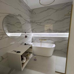 CONNECT MEBLE - Nowoczesna łazienka z marmurowymi płytkami, owalną wanną i podświetlanym lustrem. Biała szafka pod umywalką z otwartą półką. Złote detale armatury.