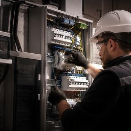 TBR RYSZARD ZWIERZYCKI - Elektryk w białym kasku i okularach, w czarnych rękawiczkach, precyzyjnie podłącza kolorowe przewody w otwartej rozdzielnicy elektrycznej, używając śrubokręta.