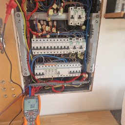 TBR RYSZARD ZWIERZYCKI - Otwarta skrzynka elektryczna z gęstwiną kolorowych przewodów i zabezpieczeń, obok miernik Sonel MPI-507 wskazujący wartości 5.0V i 0.6Ω, na drewnianym blacie.