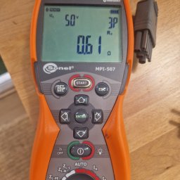 TBR RYSZARD ZWIERZYCKI - Pomarańczowo-szare urządzenie Sonel MPI-507 z wyświetlaczem pokazującym 50V i 0.61Ω, podłączone kable pomiarowe, leżące na drewnianym blacie.