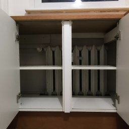 AllinOne Profesjonalnie - Biała szafka z otwartymi drzwiczkami, wewnątrz widoczny grzejnik żeberkowy. Nad szafką okno z widokiem na drzewa i niebo. Ujęcie z dołu.