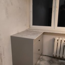 AllinOne Profesjonalnie - Szara szafka z szufladami i blatem, dopasowana do wnęki okiennej. Widoczny grzejnik i płytki podłogowe. Ściana z widocznymi śladami wilgoci.