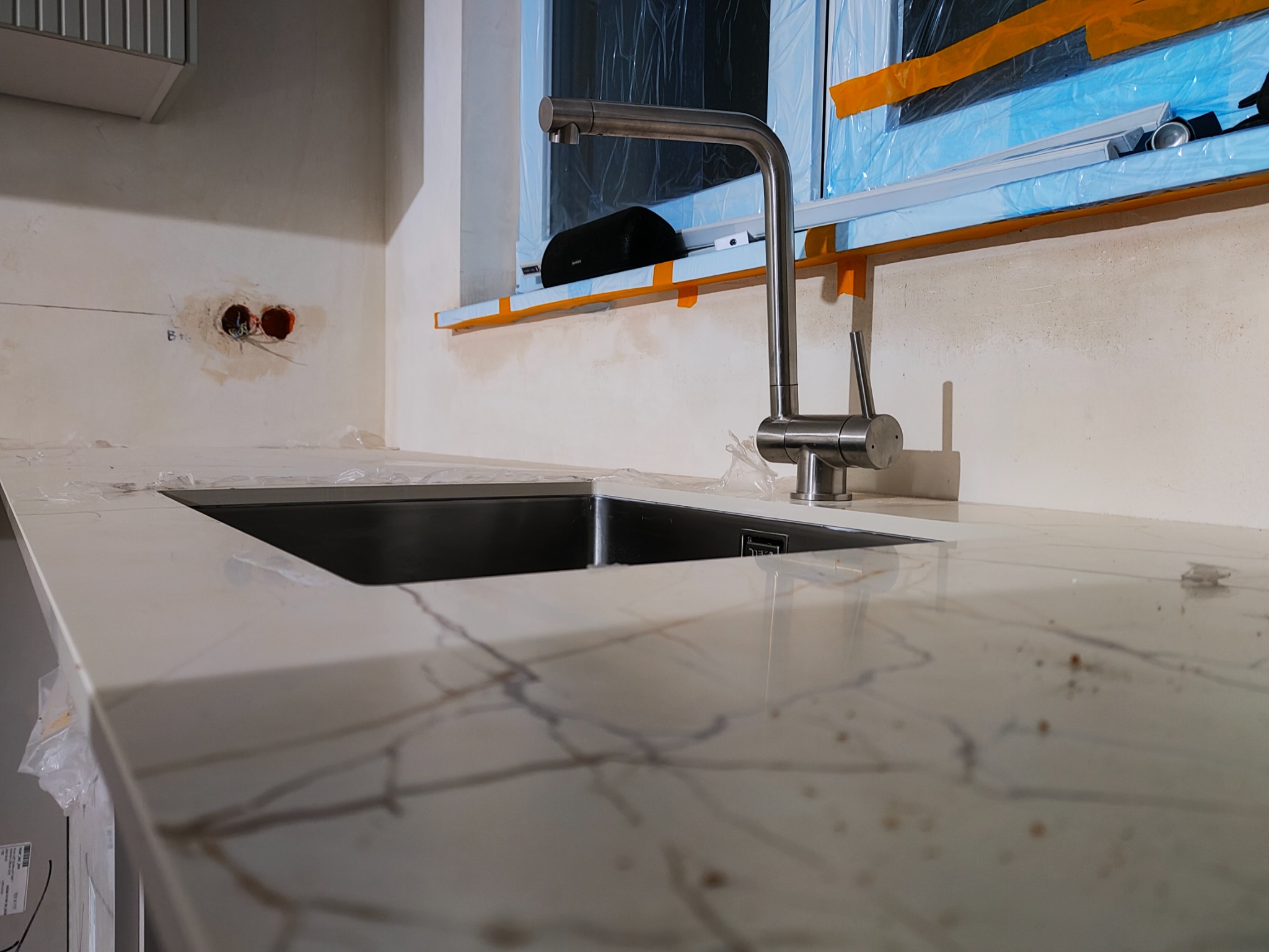 Nowoczesny blat kuchenny z marmurowym wzorem i stalową armaturą, okno zabezpieczone folią malarską. Widoczne gniazdka elektryczne w ścianie, w trakcie remontu.