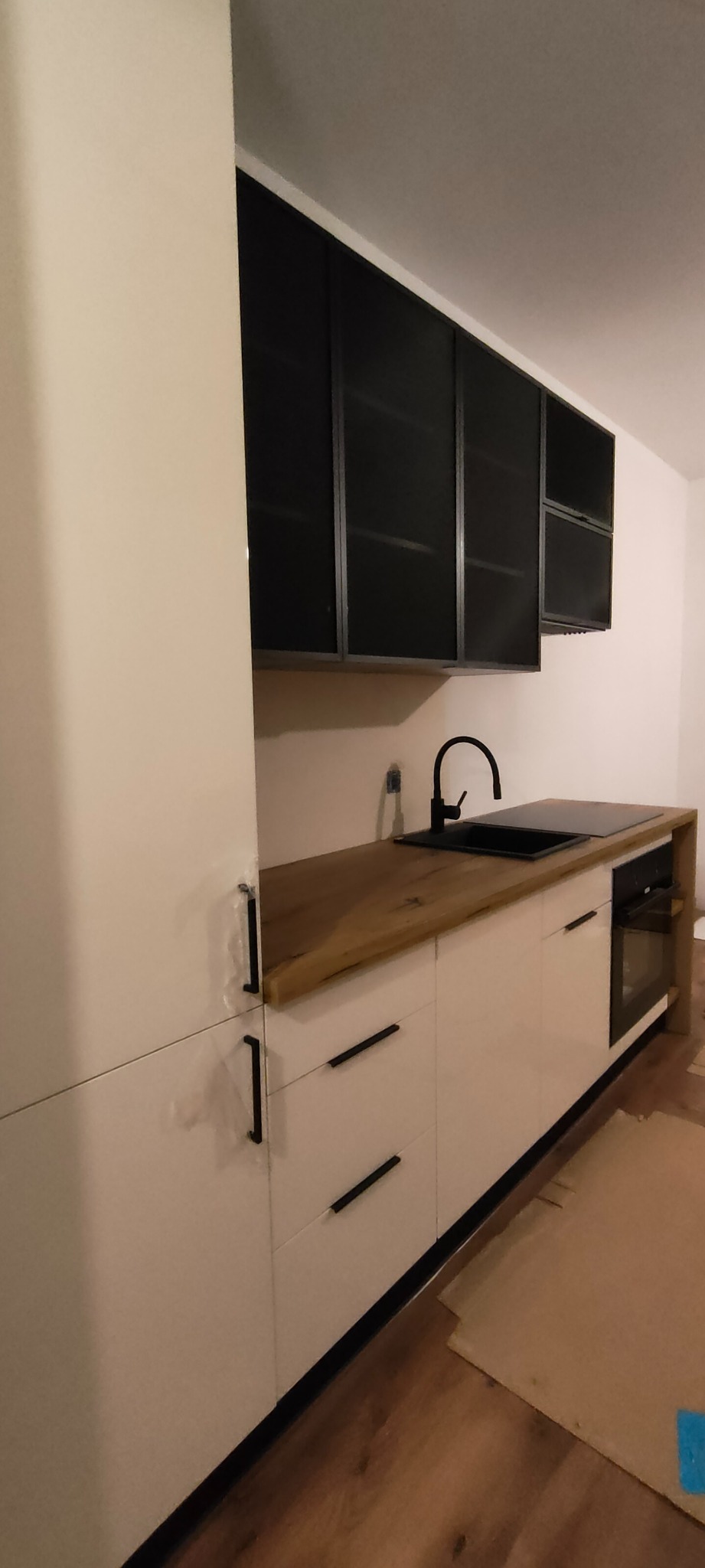 Nowoczesna kuchnia z białymi szafkami, drewnianym blatem i czarnymi akcentami: bateria, zlew, uchwyty. Górne szafki z czarnymi ramami i przeszklonymi frontami.