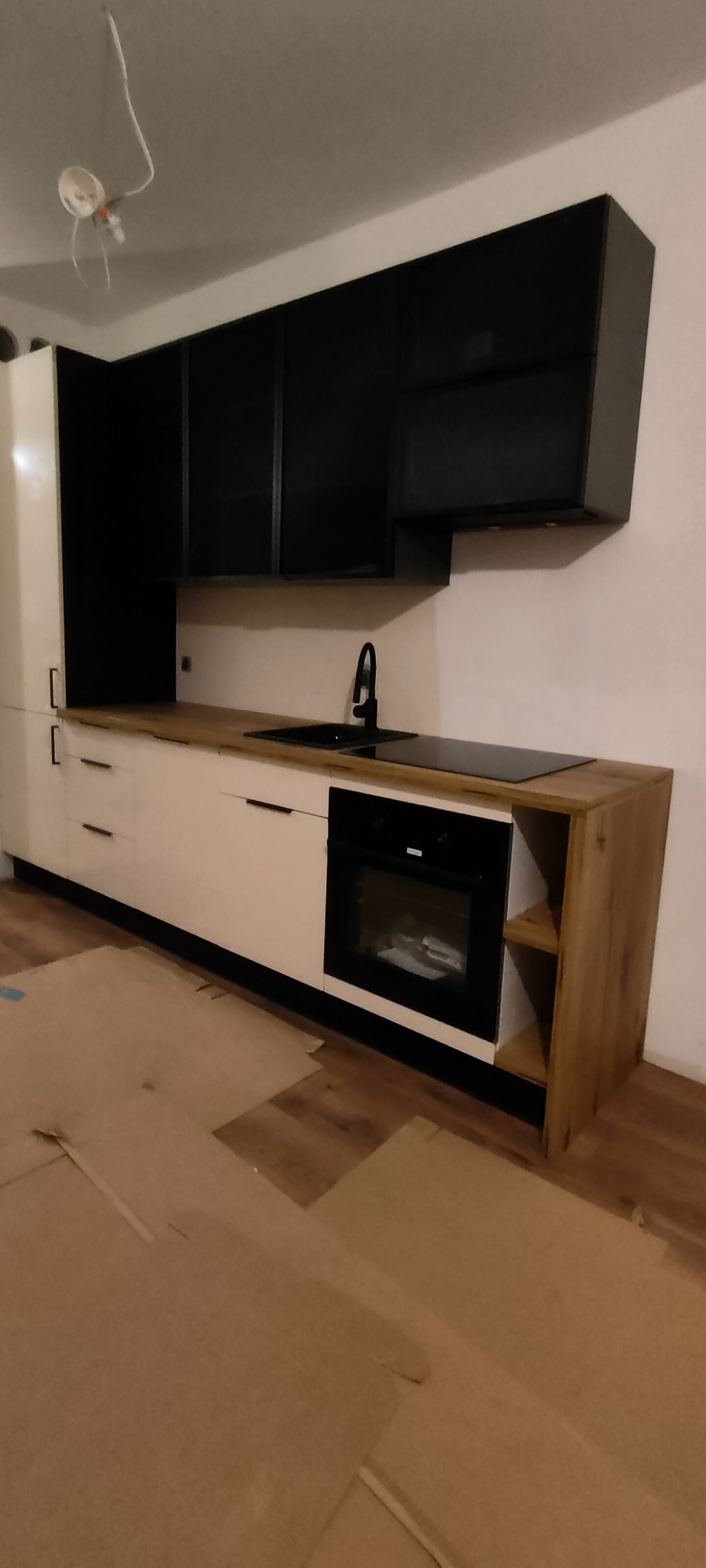 Nowoczesna kuchnia z białymi szafkami dolnymi, drewnianym blatem i czarnymi szafkami górnymi. Widoczny zlew, płyta indukcyjna i piekarnik. Podłoga zabezpieczona kartonem.