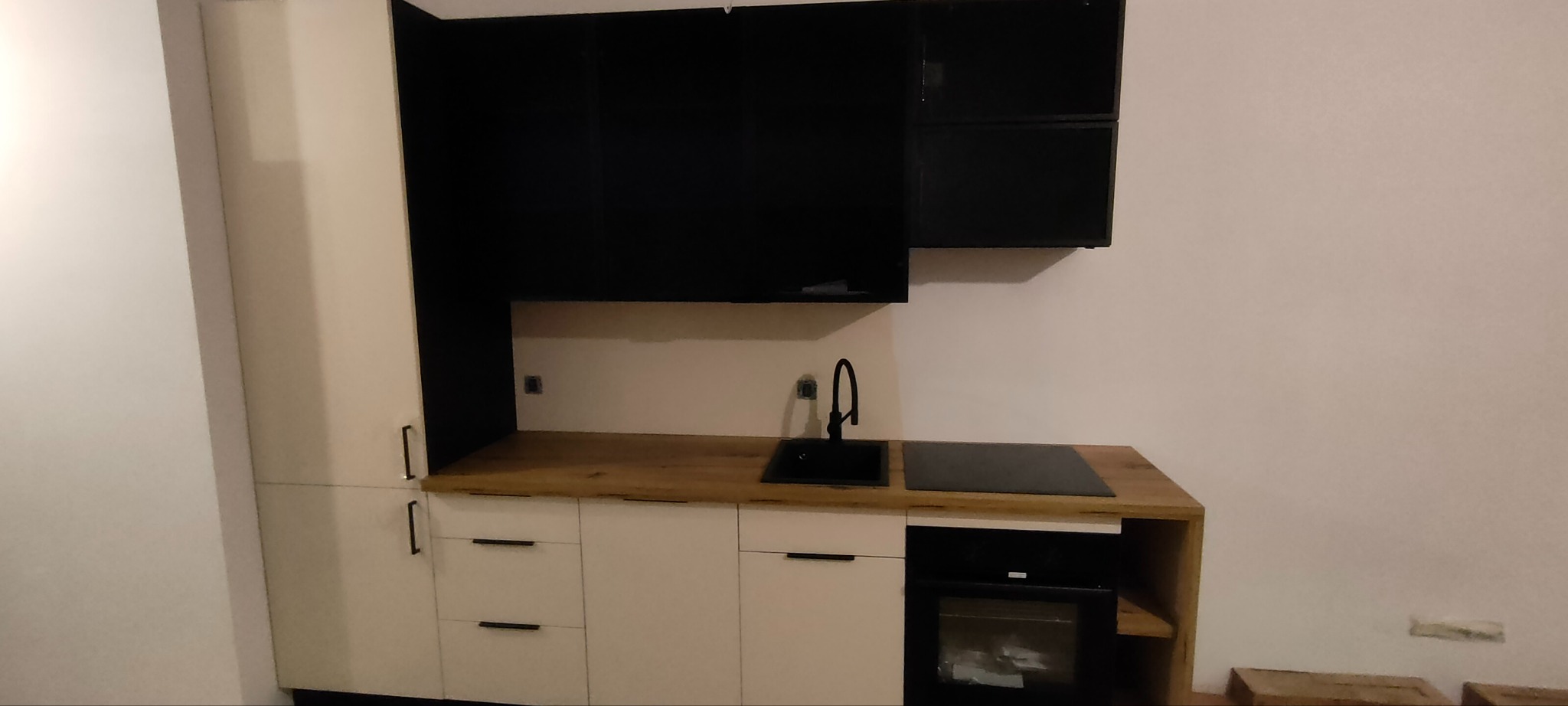 Nowoczesna, mała kuchnia z białymi szafkami dolnymi i czarnymi górnymi. Blat drewniany, czarny zlew i płyta indukcyjna. Widoczny piekarnik. Minimalistyczny design.